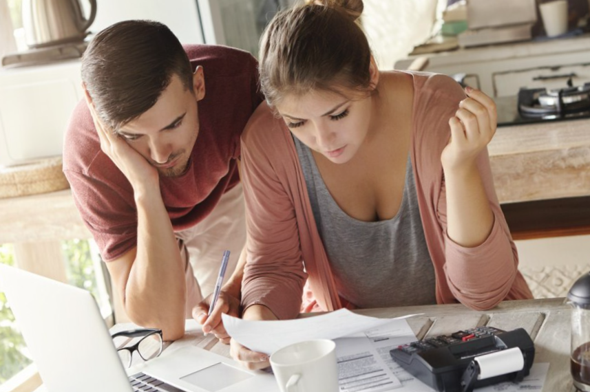 Young married couple with many debts doing paperwork together, reviewing their bills, planning family budget and calculating finances at kitchen table with papers
